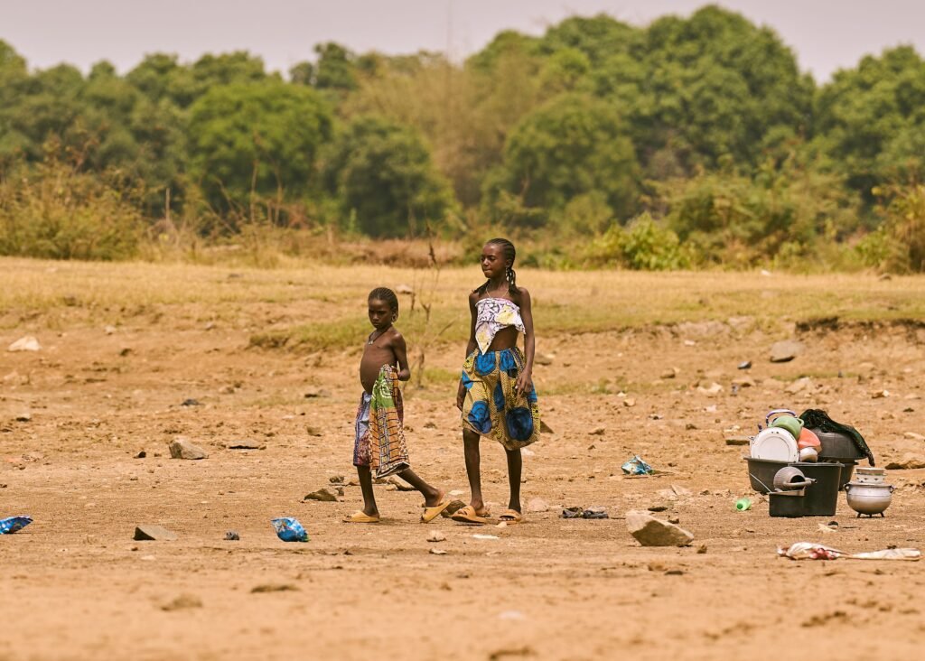 Two children stand outdoors in a rural area near Minna, Nigeria, with household items nearby.